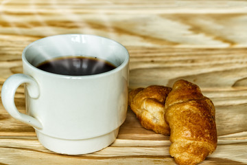 White cup with coffee and croissants on a wooden background, selective focus. The concept of breakfast.