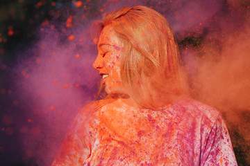 Closeup shot of a lovely caucasian blonde woman posing with exploding orange and pink Holi paint