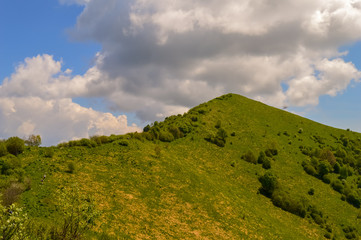 A scenic summer day view of a green mountain hill with blue sky and clouds and the peak visible