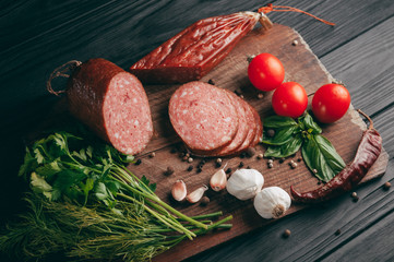 Smoked salami on a black table and a brown board. Sausages with rosemary, garlic and pepper and tomato cherry, dill, parsley.