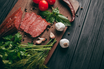 Smoked salami on a black table and a brown board. Sausages with rosemary, garlic and pepper and tomato cherry, dill, parsley.