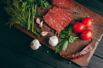 Smoked salami on a black table and a brown board. Sausages with rosemary, garlic and pepper and tomato cherry, dill, parsley.