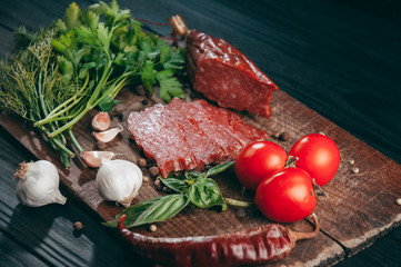 Smoked salami on a black table and a brown board. Sausages with rosemary, garlic and pepper and tomato cherry, dill, parsley