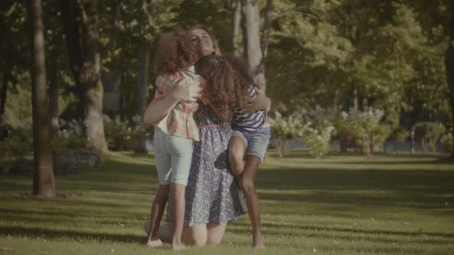 Portrait of two adorable preteen mixed race girls embracing and kissing their happy caucasian mother in summer park. Joyful multi ethnic family bonding while relaxing together in nature. Slo mo.