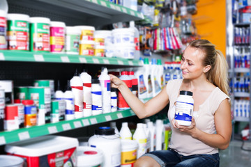smiling young woman customer choosing paint medium in bottle in hypermarket