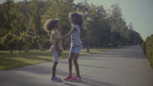 Caring cute curly preteen african american girl helping her younger sister to tie shoes while walking together in summer park. Pretty little girl fastens shoes of younger sister and embracing outdoors