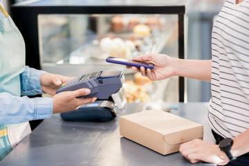 Side view mid section of unrecognizable woman paying by smartphone  via NFC for her order in shop...