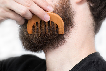 Closeup of a young man styling his long beard with a comb while standing alone in a studio against...