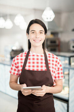 Waist up portrait of young waitress wearing apron holding note book posing looking at camera in cake shop or cafe and smiling, copy space