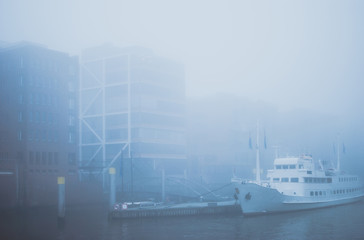 Dark gloomy cityscape of Hafen city. Gray dense fog covering the water, red bricked warehouse buildings, and boats. Obscure gothic view