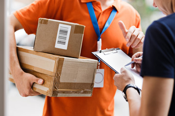 A person wearing an orange T-shirt and a name tag is delivering a parcel to a client, who is putting his signature on the receipt. Friendly worker, high quality delivery service. - obrazy, fototapety, plakaty