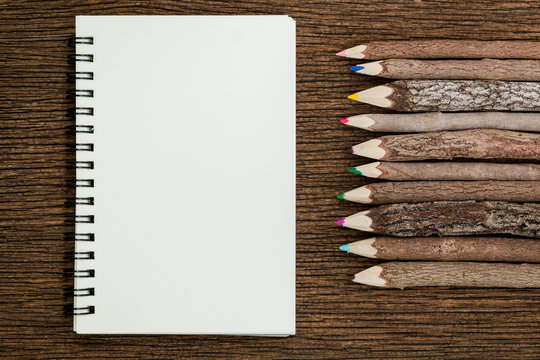 Top view of white blank empty notebook and wooden tree branch colorful pencils on wooden background.