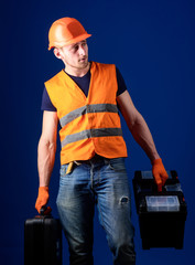 Worker, handyman, repairman, builder on calm face carries bags with professional tools. Man in helmet, hard hat holds toolbox and suitcase with tools, blue background. Equipped repairman concept.