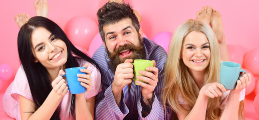 Threesome relax in morning with coffee. Lovers in bed concept. Lovers drinking coffee in bed. Man and women in domestic clothes, pajamas. Man and women, friends on happy faces lay, pink background.