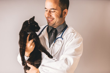 A veterinary doctor with a stethoscope around his neck holds a black cat and smiles