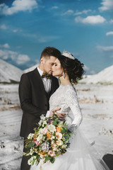 Sexy and beautiful brunette model girl with stylish hairstyle and with diadem, in white lace dress and stylish handsome men in trendy suit posing together outdoors at salts desert