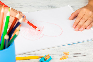 Hand of children drawing red heart with colored pencil on white paper on wooden table.