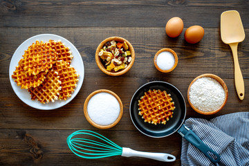 Preparing belgian waffles. Waffle on pan near whisk for whipping and ingrdients. Eggs, flour, sugar, dried fruits on dark wooden background top view