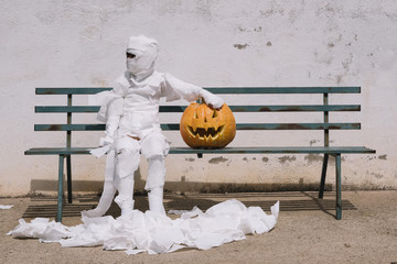 Little mummy boy with a pumpkin sitting in a street bench