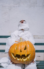 Little mummy boy with a pumpkin sitting in a street bench