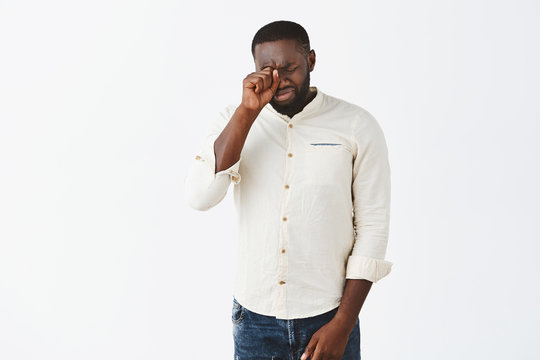 Studio shot of good-looking timid and insecure adult African American man with beard in white shirt, giving up, crying, whiping tears with fist on closed eyes, whining, having bad day over grey wall