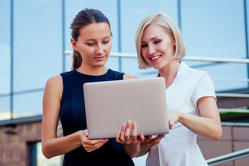 successful blonde business woman and brunette female person holding file folders and looking at securities laptop in the hands skyscraper background