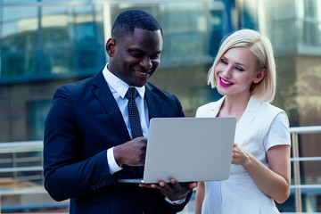 handsome African American businessman in a stylish black suit working laptop with attractive blonde woman business lady on the street background office skyscraper. successful deal idea