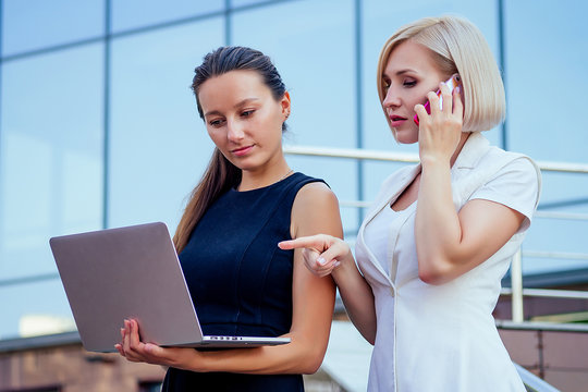 successful blonde business woman talking on the phone and brunette secretary female person holding file folders and looking at securities laptop in the hands skyscraper background