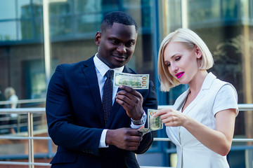 handsome Afro businessman in a stylish black suit with attractive blonde business lady consider cash money on street background office skyscraper. credit and debit idea