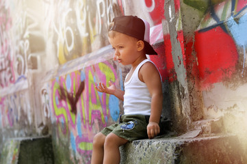 A two-year-old baby boy in a hip hop style clothes is sitting alone under the wall with graffiti and very upset