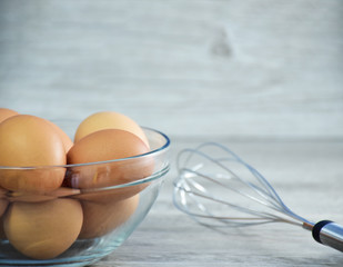 Fototapeta na wymiar Closeup of brown chicken eggs/hen eggs in a clear glass bowl with whisk on the light grey wooden background, fresh food ingredient concept, selective focus with copy space for text