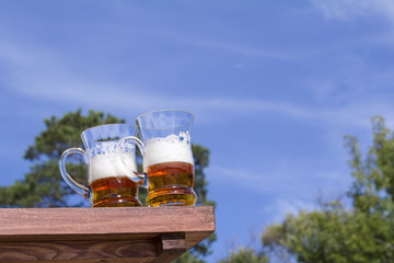Two muggs of beer are on the table against the sky and clouds