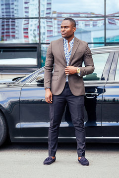 successful businessman handsome African American man in a stylish suit in brown jacket and glasses standing in front of a cool new black car on the street