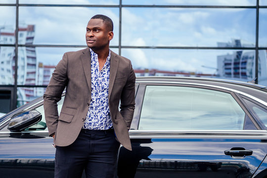 successful businessman handsome African American man in a stylish suit in brown jacket and glasses standing in front of a cool new black car on the street