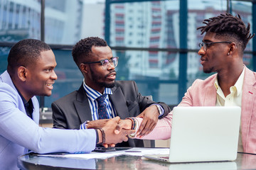 A group of three stylish African American businessman friends entrepreneurs fashion business suits meeting sitting at table and handshaking in a summer cafe outdoors. concept of successful good deal