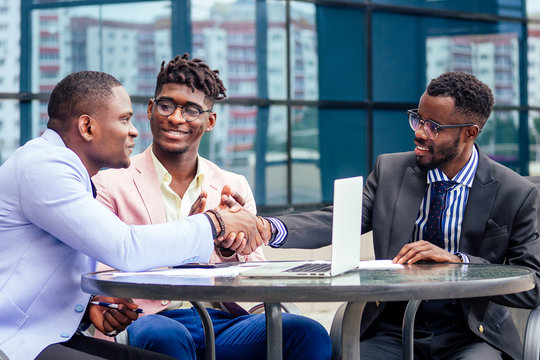 A group of three stylish African American businessman friends entrepreneurs fashion business suits meeting sitting at table and handshaking in a summer cafe outdoors. concept of successful good deal
