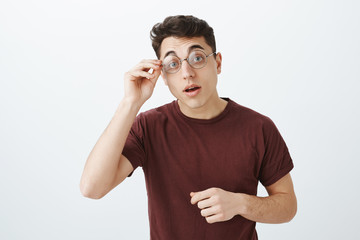 Studio shot of smart curious handsome man in round glasses and red t-shirt, raising eyebrows with...