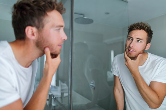 Man face care looking in mirror touching his beard or skin health. Male beauty morning skincare routine in home bathroom. After shave men lifestyle concept.