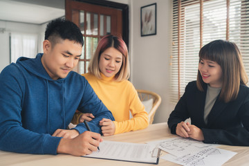 Happy young Asian couple and realtor agent. Cheerful young man signing some documents while sitting at desk together with his wife. Buying new house real estate. Signing good condition contract.