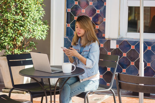 Cheerful asian young woman sitting in cafe drinking coffee and using smartphone for talking, reading and texting. Attractive asian woman holding a cup of coffee while working on laptop.