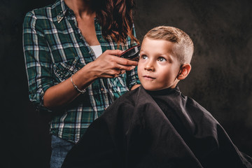 Children hairdresser with the trimmer is cutting little boy against the dark background. Cute preschooler boy getting haircut. 