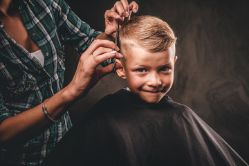Children hairdresser with scissors is cutting little boy against a dark background. Contented cute...