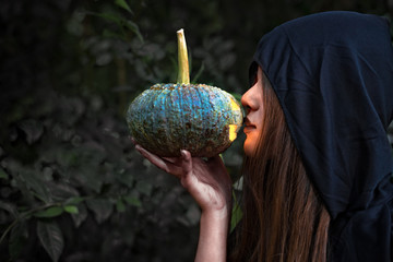 Female wizard kissing pumpkin lantern. Female costume in black clothes and hood on mystery forest background. Halloween day and Mystery concept. Fantasy of magic theme. Afterlife and Death concept.