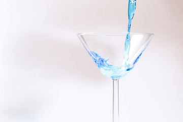 Cocktail with blue liquid in glass. Glass with blue water pouring with liquid with splashes and drops. Martini glass filling with alcohol with splashes on white background. Refreshing drink concept.