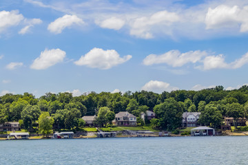 Naklejka na ściany i meble Upscale homes with boat docks built along the edge of a lake with tall green trees under a blue sky with fluffy clouds