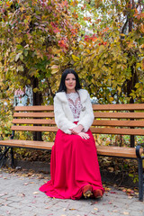 Attractive brunette woman in red dress sitting on bench in autumn park