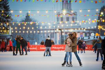 Young couple in love Caucasian man with blond hair with long hair and beard and beautiful woman have fun, active date skating on ice scene in town square in winter on Christmas Eve