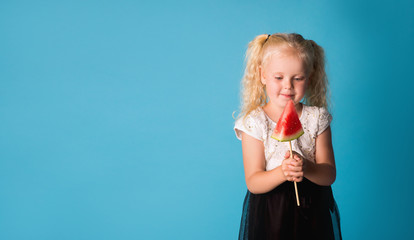 Portrait of a young girl with long hair looking at a delicious piece of watermelon on a stick in her hand with eyes and tongue out isolated on a blue background