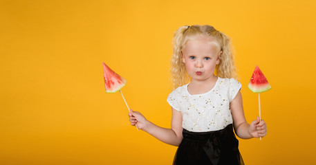 Portrait of a young girl with long hair looking at a delicious piece of watermelon on a stick in the hand with eyes and tongue out isolated on a yellow background