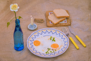 Fried eggs on the ornamental plate. Yellow rose in the blue bottle, blue candlestick with burning candle, yellow fork and knife, 2 slices of bread on the rough table cloth.